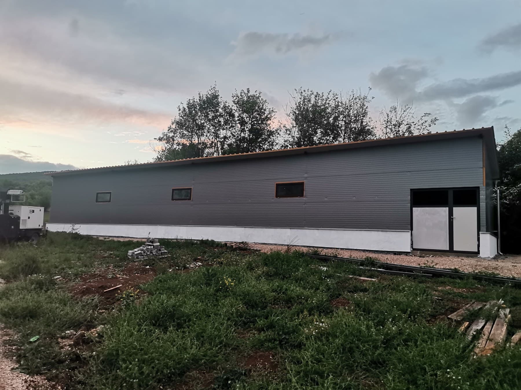Dugačak objekat za živinu od sivih panela u sumrak, okružen zelenilom